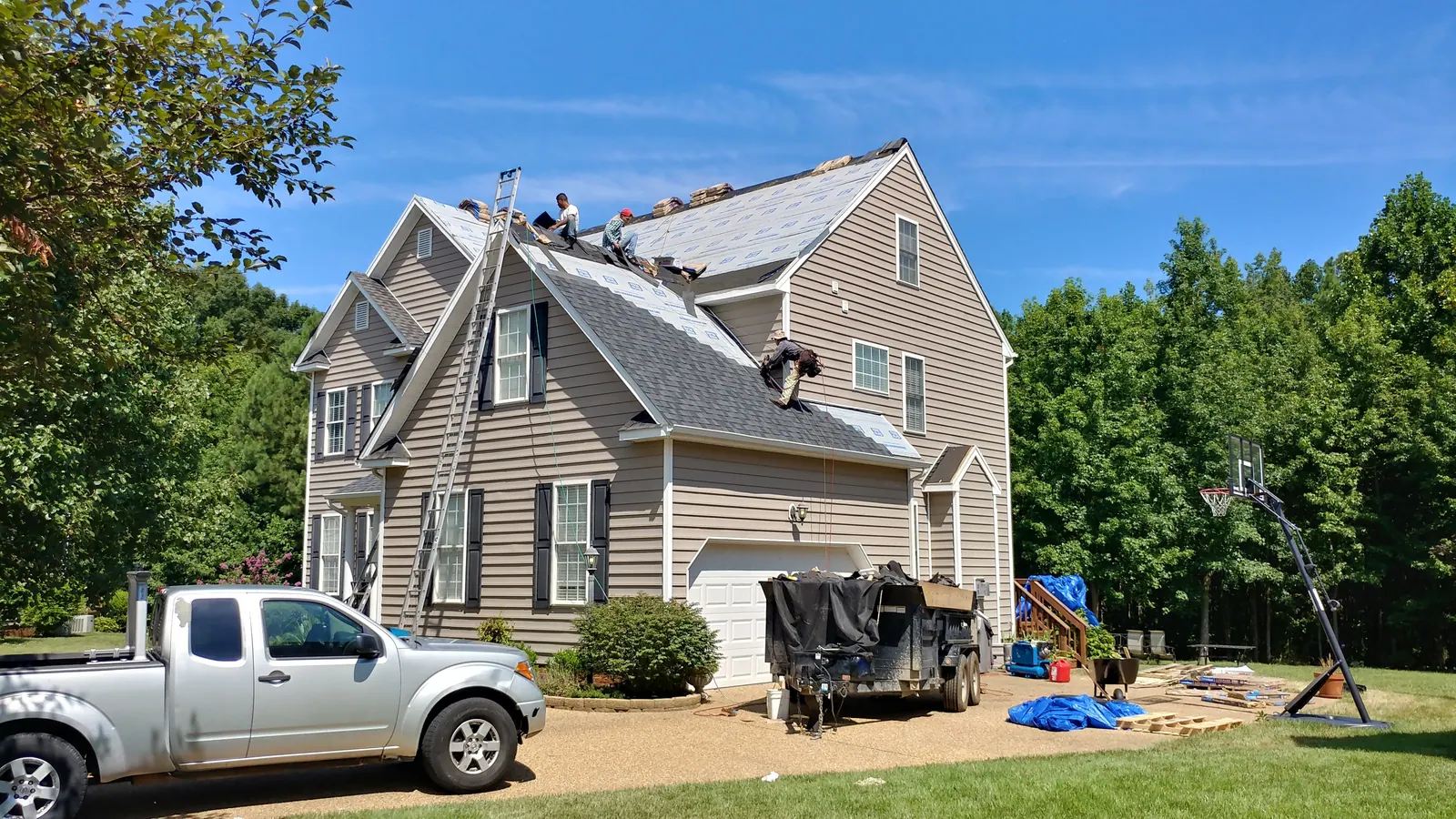 Crew replacing shingles on a steep-pitch roof with a dump trailer in the driveway